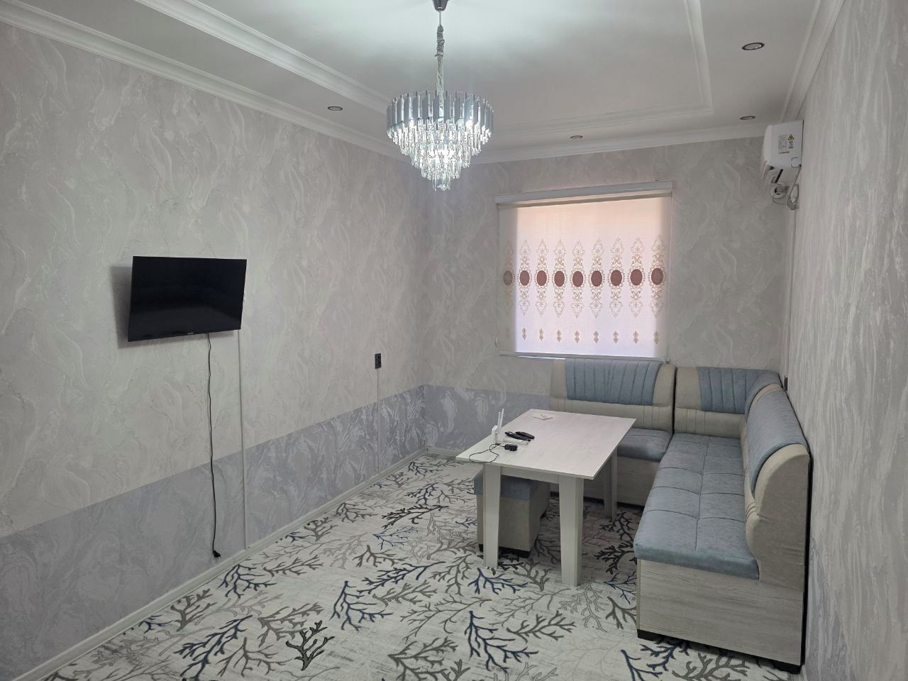 A room with a television, a table, a sofa, a chandelier, and a patterned carpet.