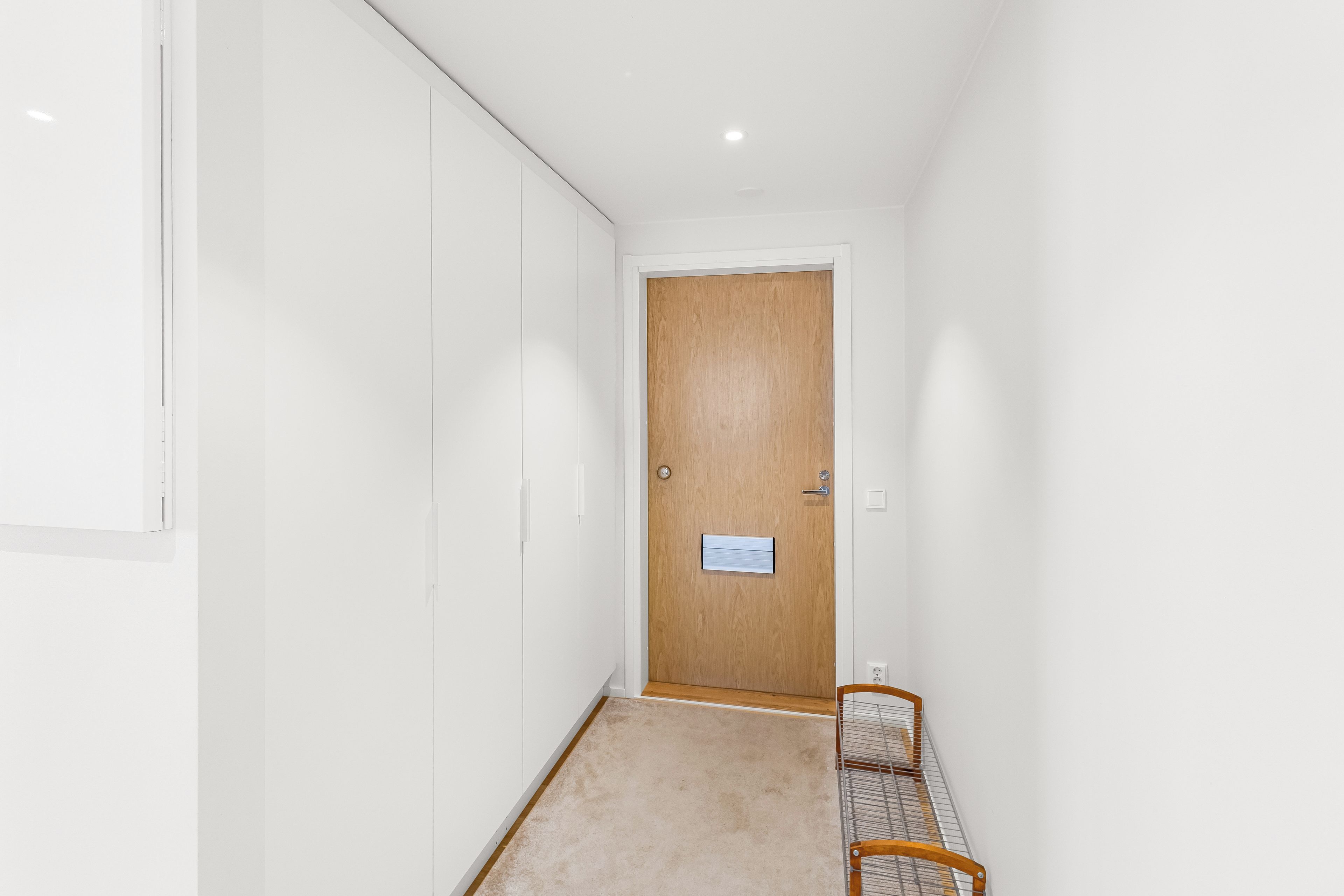 Un couloir étroit avec une porte en bois, un banc et un mur blanc.