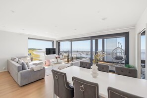 Ett modernt vardagsrum med en soffa, ett matbord och stolar, stora fönster med utsikt över havet och en balkong med utemöbler.