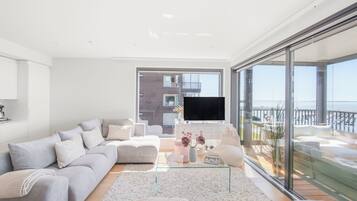A modern living room with a large sofa, a coffee table, and a TV. There is a balcony with a view of the sea.