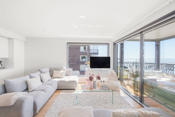 A modern living room with a large sofa, a coffee table, and a TV. There is a balcony with a view of the sea.