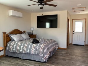 A bedroom with a bed, a ceiling fan, a mounted TV, a bedside table with a lamp, and a door. - Ben's Place - A Quiet Sanctuary where you can enjoy sunrises and sunsets. (Sonoita)