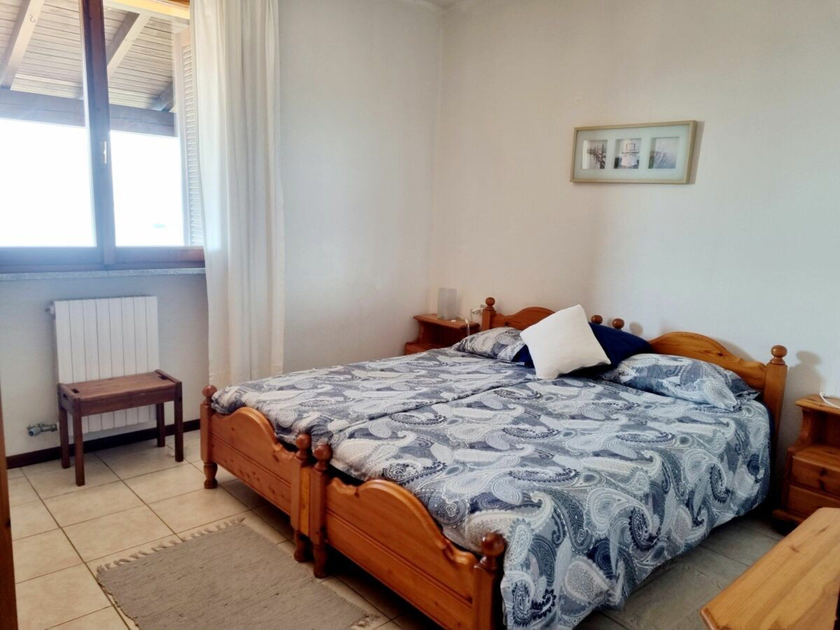 Une chambre à coucher avec un lit en bois, une table de chevet, une commode, un petit tabouret en bois et un radiateur.
