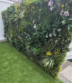 Eine vertikale Begrünung mit verschiedenen Pflanzen und Blumen, eine Rasenfläche im Vordergrund und eine weiße Wand im Hintergrund.