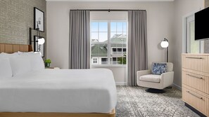 Ein Hotelzimmer mit einem großen Bett, einem Sessel, einem Schreibtisch, einer Lampe, einem Fernseher und einem Fenster mit Vorhängen.