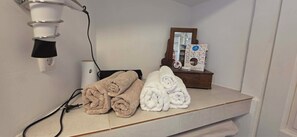 Un baño con un perchero que sostiene toallas enrolladas, un secador de pelo y un pequeño cofre de madera con un espejo y una caja de pañuelos.