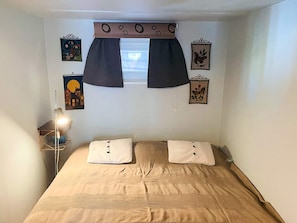 Un dormitorio con cama, dos almohadas, una mesita de noche con lámpara, una ventana con cortinas y adornos en la pared.