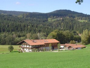 A two-story building with a red-tiled roof, surrounded by a green field and forested hills. - Starry sky - Groller Holiday Farm (Drachselsried)