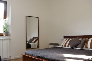 A bedroom with a bed, a bedside table, a mirror, a radiator, and a window with a plant.