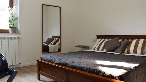 A bedroom with a bed, a bedside table, a chair, a radiator, a window with a plant, and a mirror. - Fly&Rest MXP | Unit 2 | 24/7 Airport Transfer (Casorate Sempione)