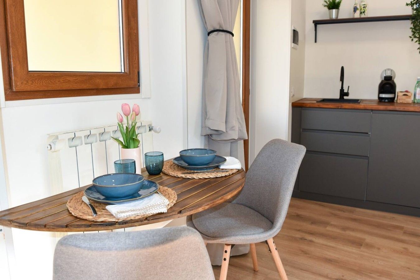 Een eethoek met een houten tafel, twee blauwe kommen, een vaas met roze bloemen en een grijze stoel.