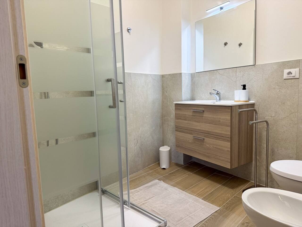 Un bagno moderno con box doccia in vetro, mobile lavabo in legno, lavandino bianco e uno specchio grande.