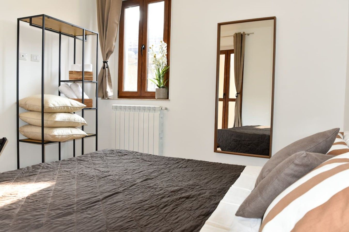 Ein Schlafzimmer mit Bett, Schrank, Heizkörper, einem Fenster mit Vorhängen und einem Spiegel.