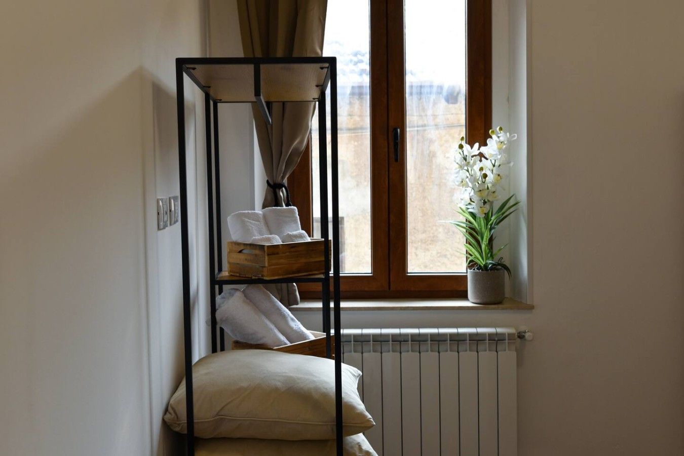 Une pièce d’angle avec un radiateur, une fenêtre avec des rideaux, une plante et une étagère en métal où sont posées des serviettes.