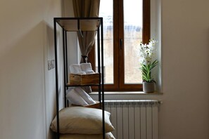 A corner room with a radiator, a window with curtains, a plant, and a metal shelf with towels.