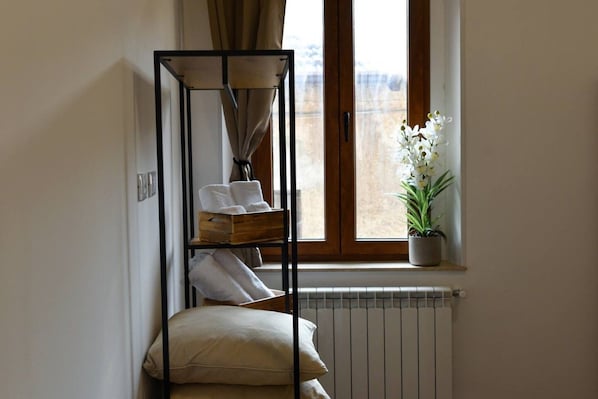 A corner room with a radiator, a window with curtains, a plant, and a metal shelf with towels.