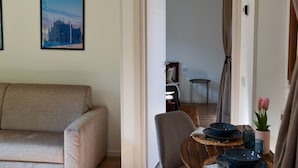A living room with a sofa, a dining table with plates and a vase of flowers, and a poster of a building on the wall. - Fly&Rest MXP | Unit 2 | 24/7 Airport Transfer (Casorate Sempione)