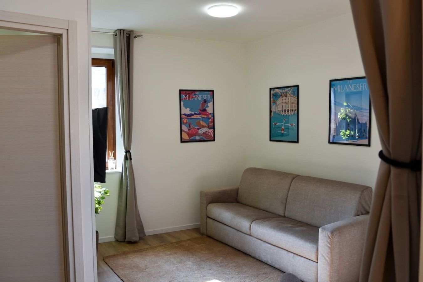 Ein Zimmer mit einer beigen Couch, einer Tür und drei gerahmten Postern an der Wand.