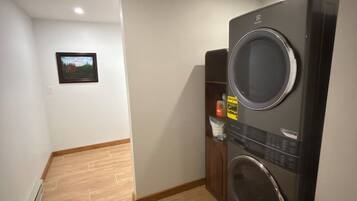 A laundry room with a front-loading washer and dryer, wooden flooring, and a small shelf with items.