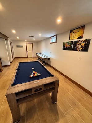 A pool table with balls, a dartboard, and wall art in a room with wooden flooring and recessed ceiling lights.