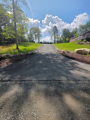 A paved road flanked by trees and a building with a sloped roof. - The 48 Loon`s Landing (Lake Placid)