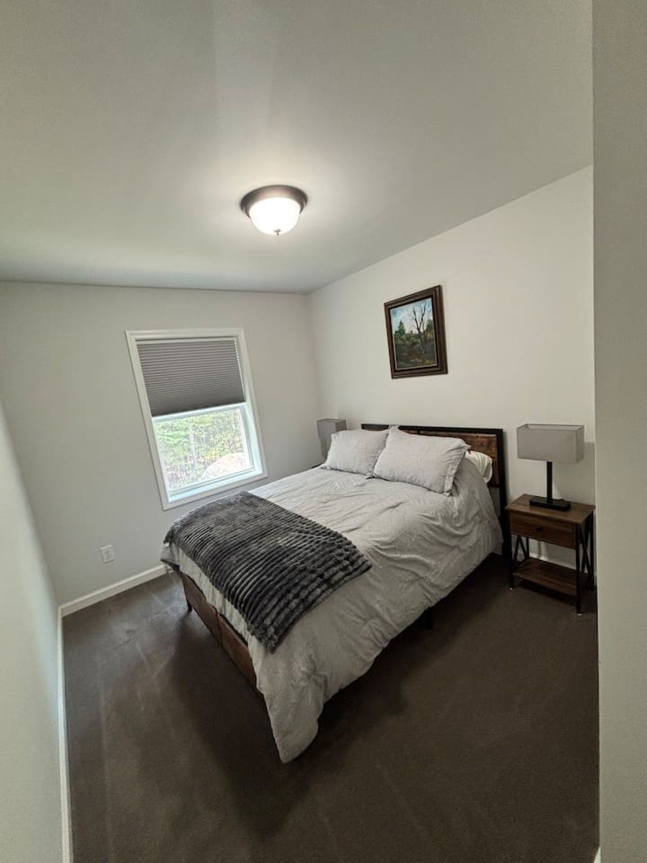 A bedroom with a bed, a bedside table with a lamp, a window with blinds, and a ceiling light.