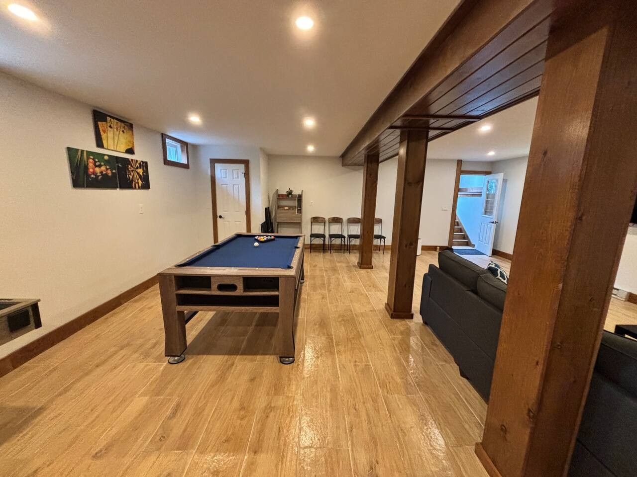 Una mesa de billar en una habitación con suelo de madera, un sofá y una escalera que lleva a otra zona.