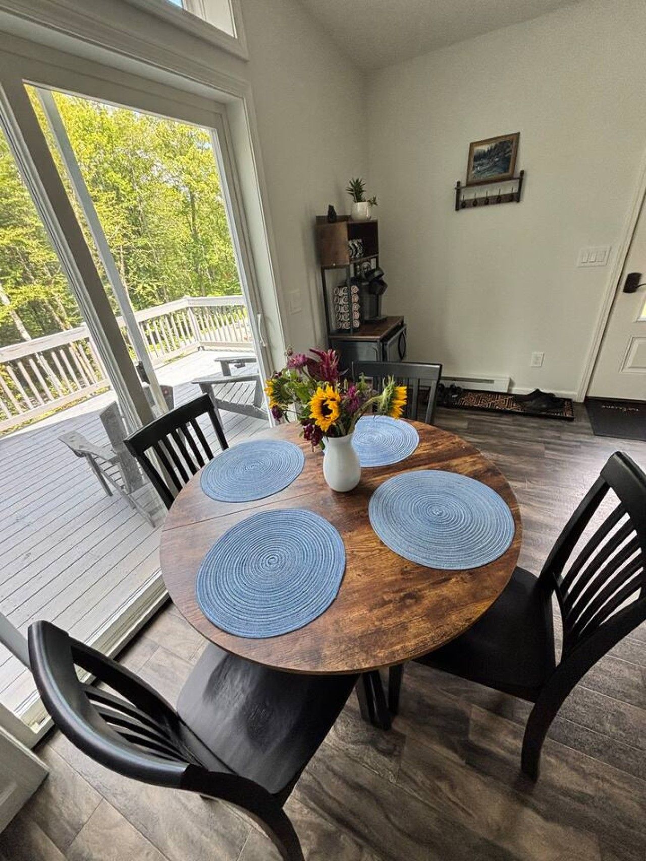 A wooden dining table with four blue placemats, surrounded by black chairs, in a room with a view of greenery through a glass door.