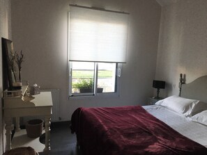 A bedroom with a bed, a window with a view, a bedside table, and a lamp. - Michelle's House (Pont-l'Abbé)
