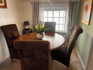 A dining area with a round table, four chairs, a vase with flowers, a lamp, and a window with blinds. - Old Cottages: 3 Bed (Ripon)