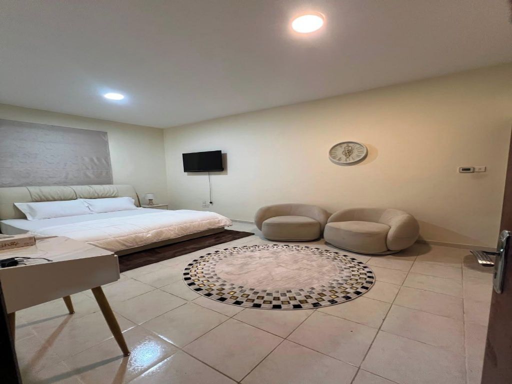 A bedroom with a bed, a desk, a chair, a round rug, a television, and a clock on the wall.