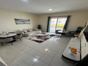 A living room with a sofa, coffee table, and chairs, a dining area with a table and chairs, a large window with curtains, and a painting on the wall. - Cozy 1-bedroom apartment in peaceful Dubai with AC (Dubai)