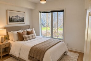 Un dormitorio con una cama grande, una mesita de noche, una lámpara, un cuadro enmarcado y una ventana que ofrece vistas a árboles y montañas.