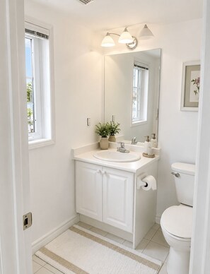 Casa de banho com lavatório branco, espelho e sanita. Há uma janela a fornecer luz natural e uma pequena planta no lavatório.