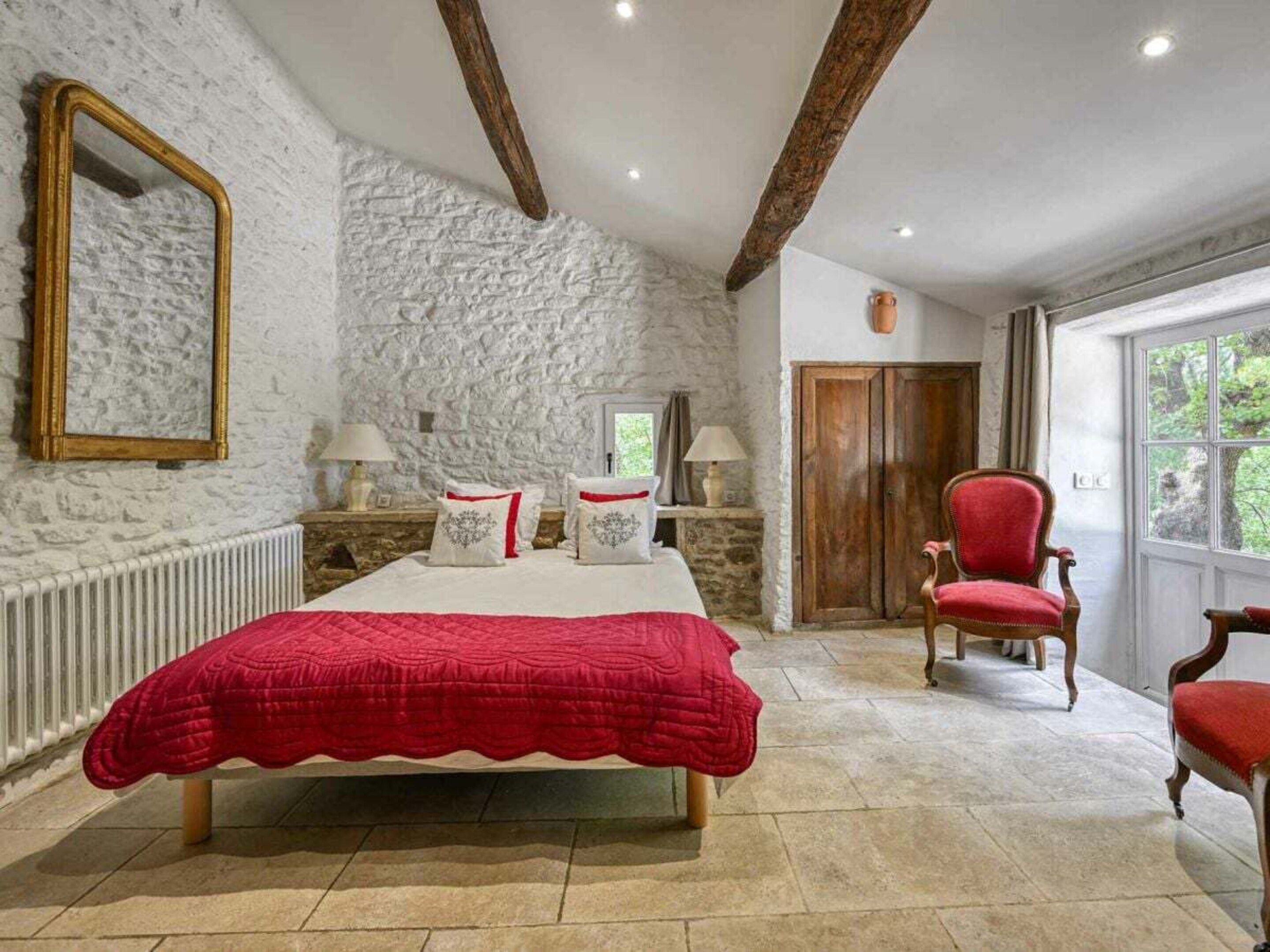 A bedroom with a stone wall, a bed with a red bedspread, two bedside lamps, a wooden chair, and a large window.
