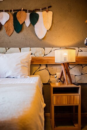 A bedroom with a stone wall, a wooden bedside table with a lamp, a bed with white bedding, and a wall hanging of woven leaves. - Casa Linda a poucos passos do mar (Sao Miguel dos Milagres)
