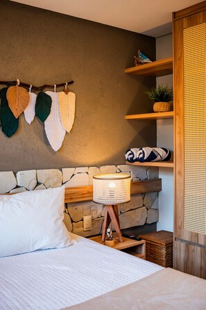 A bedroom with a stone wall, a bed with white bedding, a wooden headboard, a wall-mounted lamp, and a shelf with decorative items. - Casa Linda a poucos passos do mar (Sao Miguel dos Milagres)