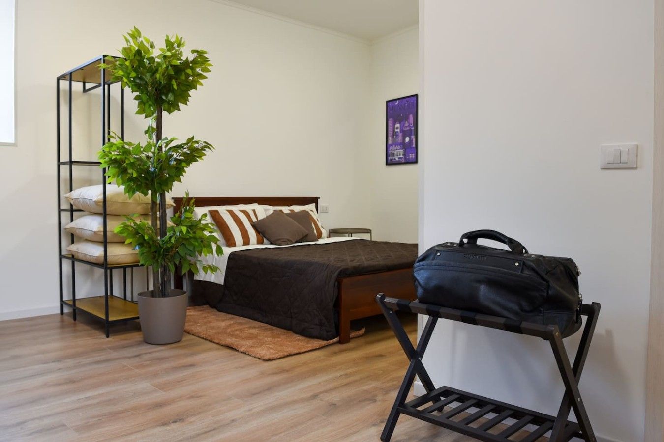 A bedroom with a bed, a potted plant, a suitcase, and a wall-mounted shelf.