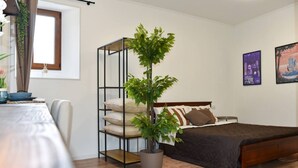 A bedroom with a bed, a bedside table, a potted plant, a window, and wall art. - Fly&Rest MXP | Suite | Airport Transfer 24/7 (Casorate Sempione)