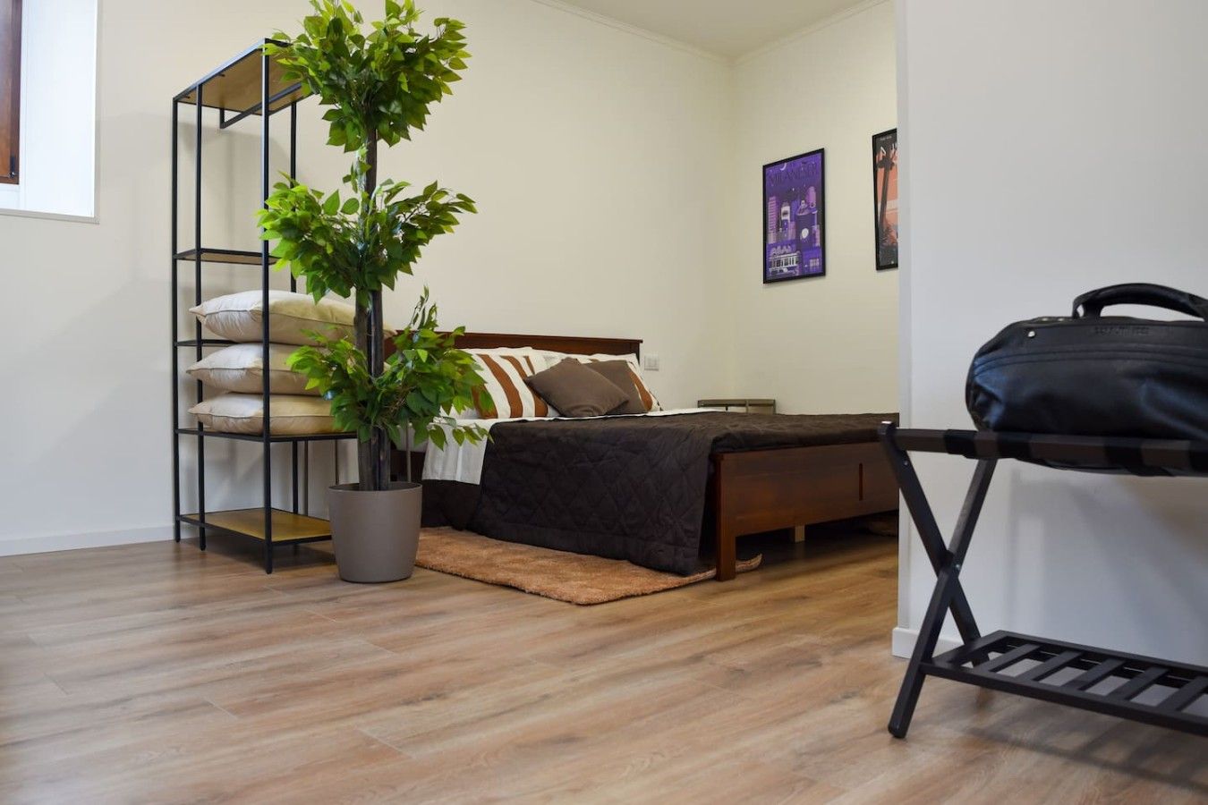Ein Schlafzimmer mit einem Bett, einer Topfpflanze, einem Regal mit Kissen und einem Koffer auf einem Beistelltisch.