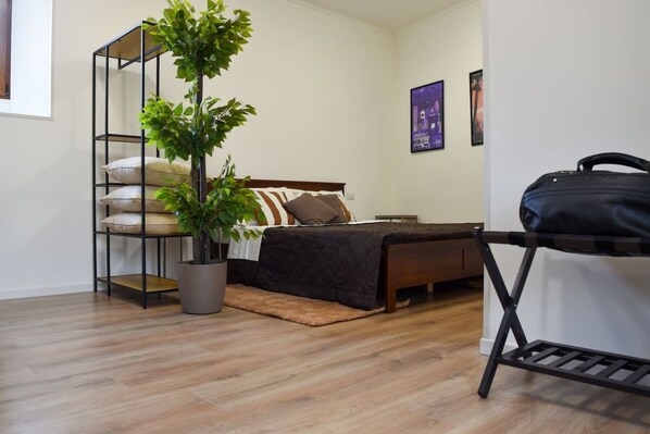 A bedroom with a bed, a potted plant, a shelf with cushions, and a suitcase on a stand.