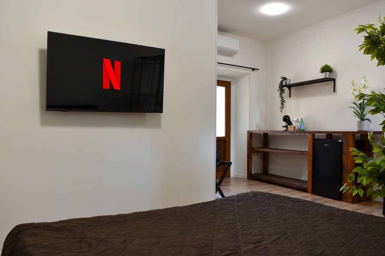 Une chambre avec un lit, un téléviseur fixé au mur affichant le logo de Netflix, une petite étagère avec une plante et un réfrigérateur.