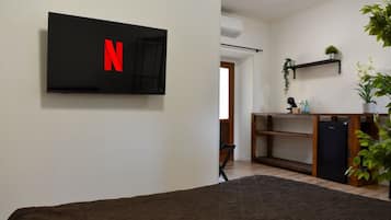 Une chambre avec un lit, un téléviseur fixé au mur affichant le logo de Netflix, une petite étagère avec une plante et un réfrigérateur.