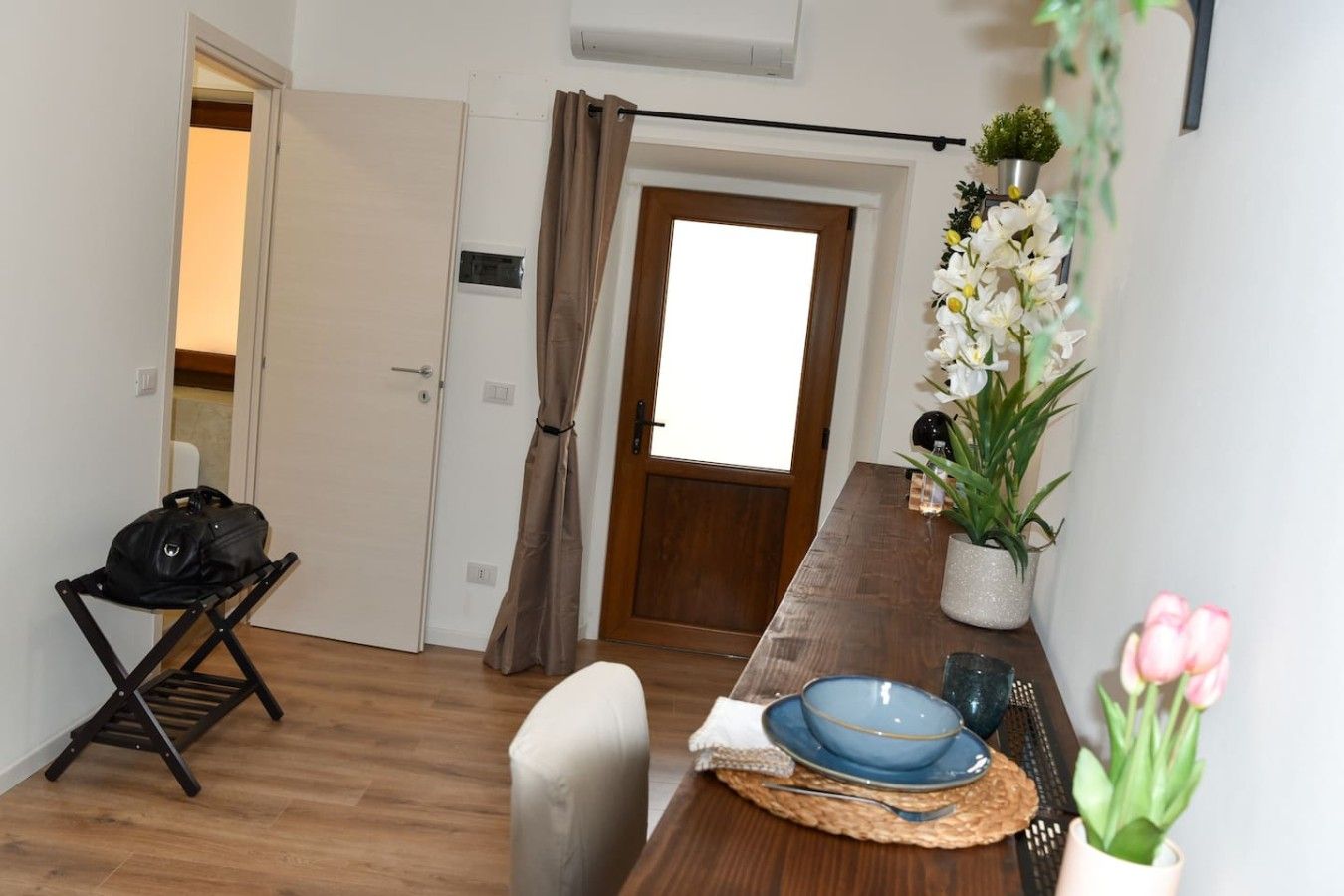 Une pièce avec un plancher en bois, une table à manger avec des assiettes bleues, un sac noir posé sur une chaise, une plante en pot et une porte avec un rideau.