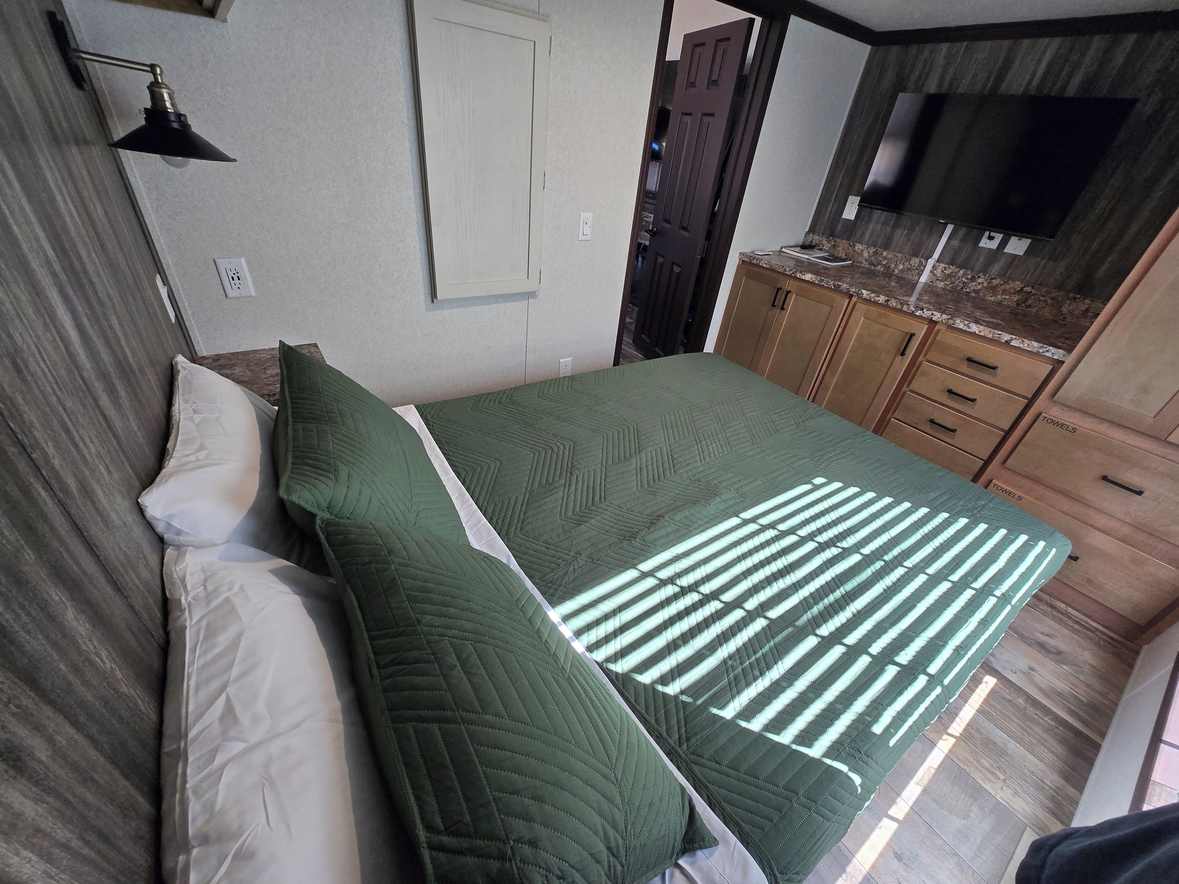 A bedroom with a bed, a nightstand, a wall-mounted lamp, a door, and a TV on a wooden cabinet.