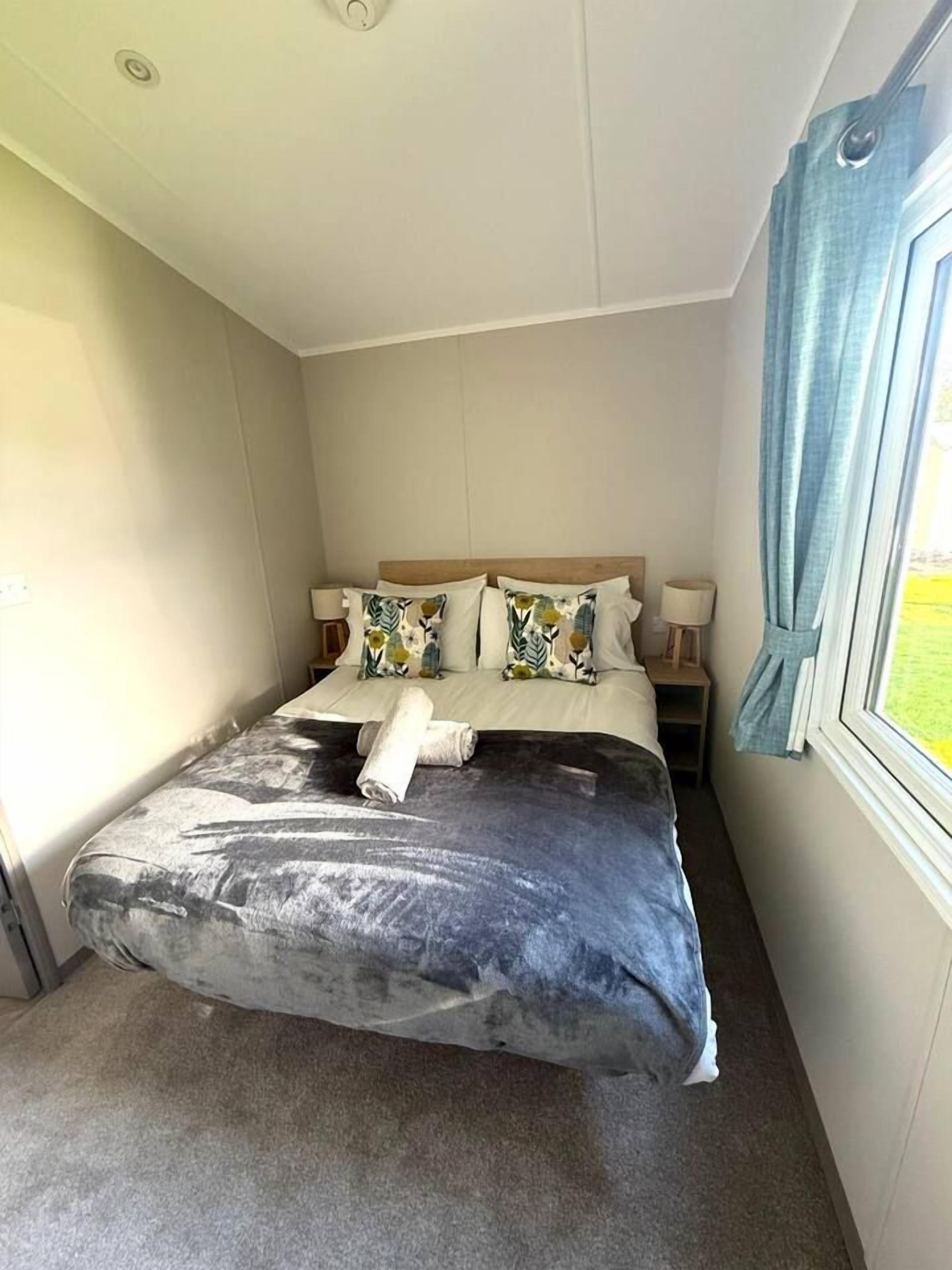 A single bed with a grey blanket and two pillows, a bedside table with a lamp, and a window with curtains.