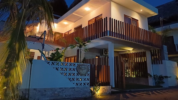 Un bâtiment moderne à deux étages, doté d’un balcon, d’une porte décorative et, en arrière-plan, d’une structure à toit de chaume.