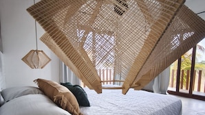 Un lit avec un cadre en bois, un luminaire suspendu en fibres tress ées et une vue sur des palmiers par la fenêtre.