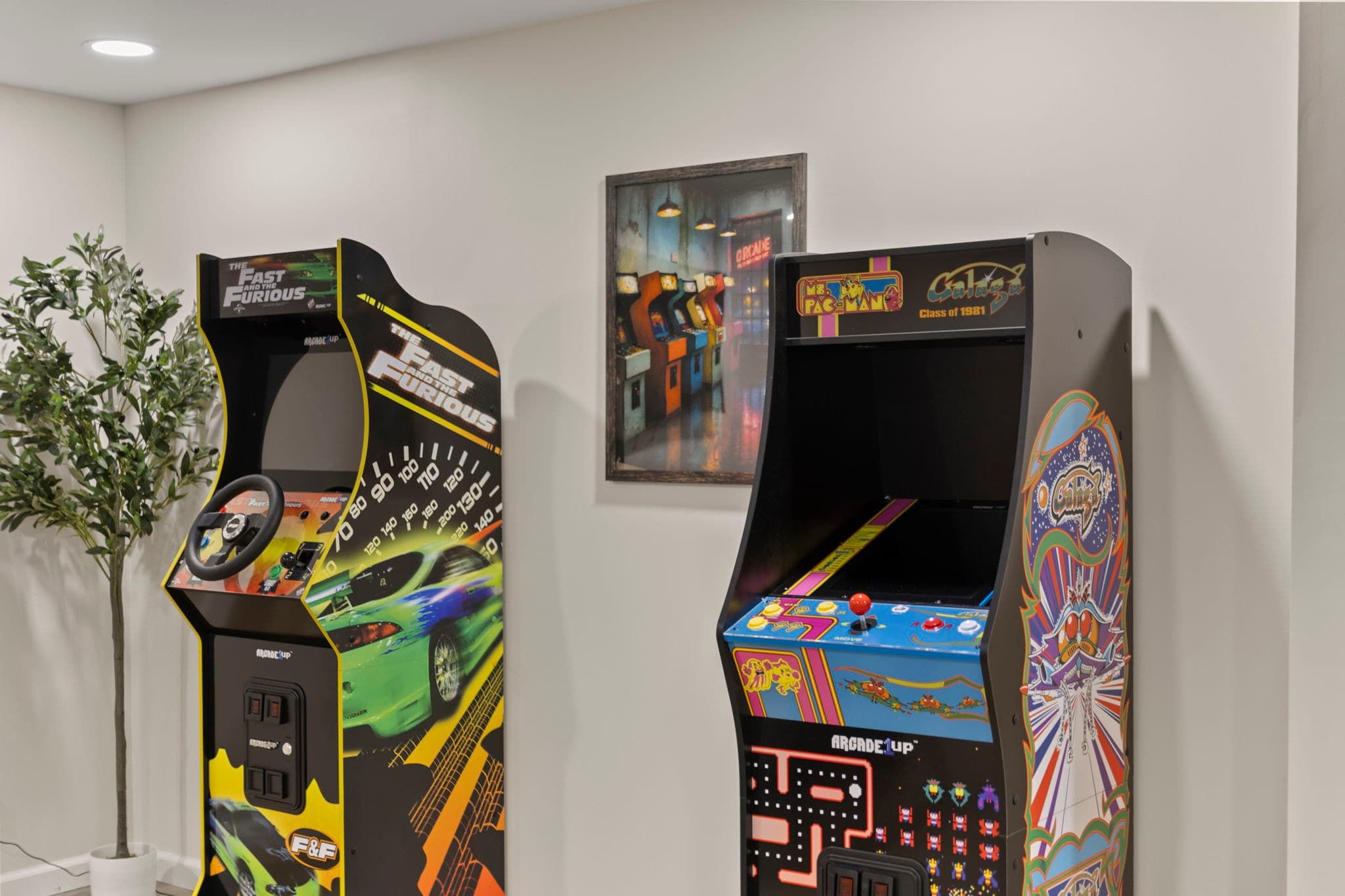 Two arcade machines with colorful graphics and controls, placed in a room with a white wall and a framed picture above them.