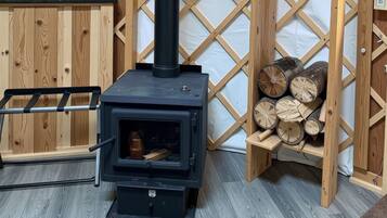 A wood-burning stove with a chimney, a metal ladder, and a stack of firewood against a wooden lattice wall.